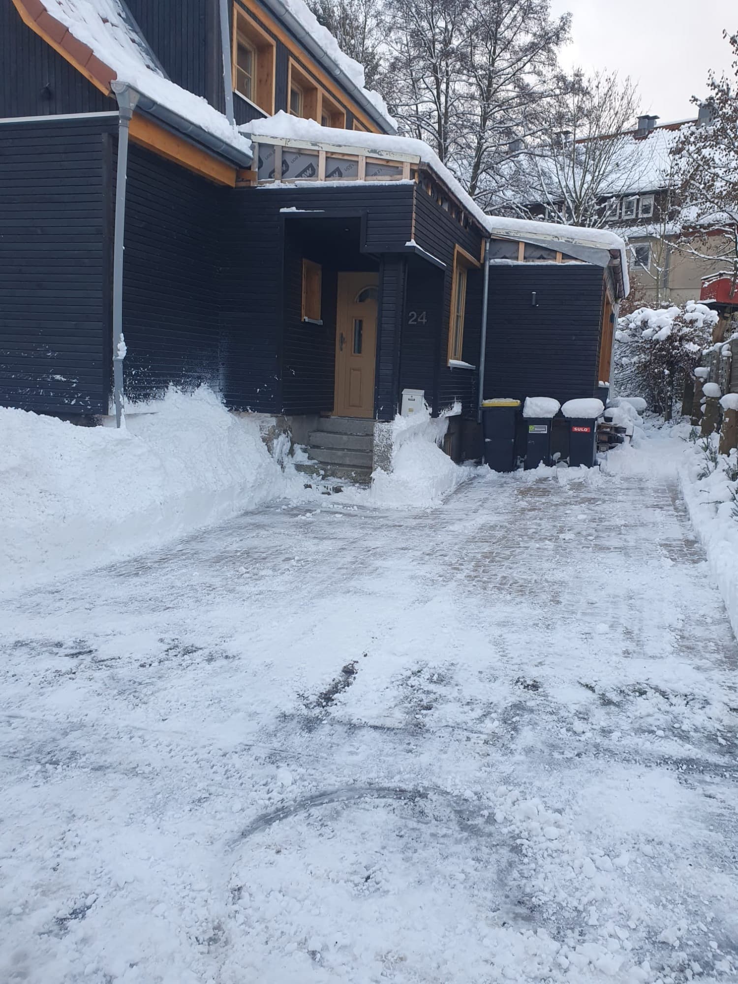 Geräumte und gestreute Hofeinfahrt vor einem dunkel verschalten Harzer Holzhaus, links und rechts hoch aufgeschobener Schnee.